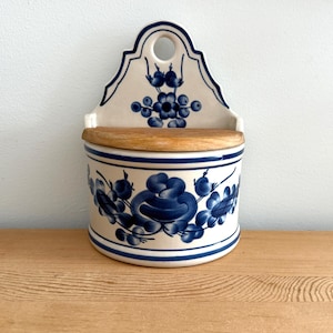 Pode incluir: Uma caixa de sal de cerâmica para parede com base branca, padrões florais azuis e tampa de madeira. O recipiente possui um painel traseiro curvo com um orifício circular para pendurar e um corpo cilíndrico decorado com flores azuis pintadas à mão.