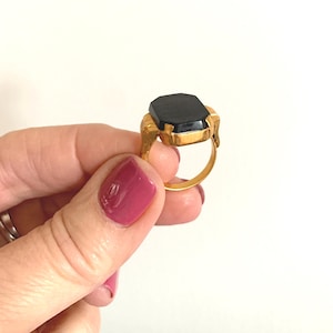 Puede incluir: Un anillo de oro con una piedra preciosa negra. El anillo tiene un engaste cuadrado con un borde biselado.