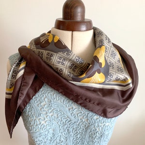 May include: A brown silk scarf with a geometric pattern in shades of beige, yellow, and brown. The scarf is tied around a mannequin's neck.