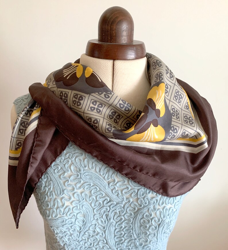 May include: A brown silk scarf with a geometric pattern in shades of beige, yellow, and brown. The scarf is tied around a mannequin's neck.