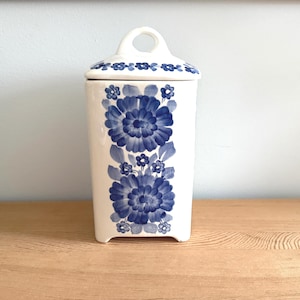 Könnte beinhalten: Ein weißer Keramikbehälter mit Deckel, verziert mit einem blauen Blumenmuster. Der Behälter hat eine rechteckige Form mit abgerundeten Ecken und einem Griff am Deckel. Das Blumenmuster umfasst große und kleine Blüten und Blätter.