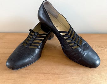 May include: A pair of black leather shoes with a low heel. The shoes have a unique design with multiple elastic straps across the top of the foot. The shoes are displayed on a wooden surface.