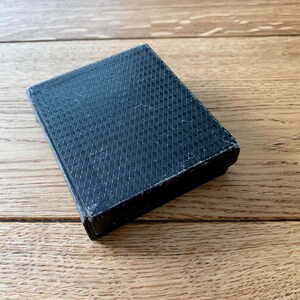 May include: A small, rectangular, black box with a textured, crosshatch pattern on the top. The box is closed and appears to be made of a fabric-like material. The box is resting on a wooden surface.
