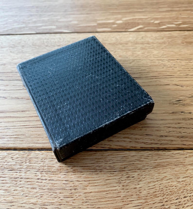 May include: A small, rectangular, black box with a textured, crosshatch pattern on the top. The box is closed and appears to be made of a fabric-like material. The box is resting on a wooden surface.