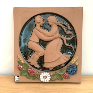 Puede incluir: Un azulejo de cerámica marrón con un diseño en relieve de una pareja bailando. La pareja está en un círculo con un fondo azul oscuro. El azulejo está decorado con un borde floral de flores rojas, blancas y azules. El azulejo está firmado "JIE" en la esquina inferior izquierda.
