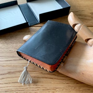 May include: A small, black leather-bound book with a decorative tassel. The book has a wooden edge and is resting on a wooden surface. An open black box is in the background.