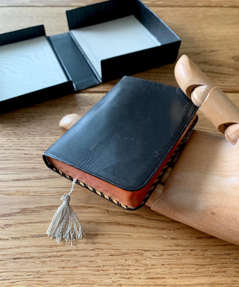 May include: A small, black leather-bound book with a decorative tassel. The book has a wooden edge and is resting on a wooden surface. An open black box is in the background.