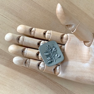 May include: A silver metal leaf-shaped stamp with a detailed design. The stamp is resting on a wooden hand model.