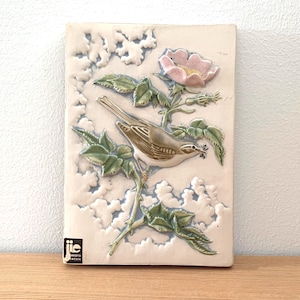 以下が含まれることがあります： 花咲く枝に止まる鳥のレリーフが施された長方形のセラミックウォールプラーク。鳥は茶色と白で、くちばしに小枝を持っています。花はピンク色で、葉は緑色です。背景はオフホワイトで雲のような模様です。