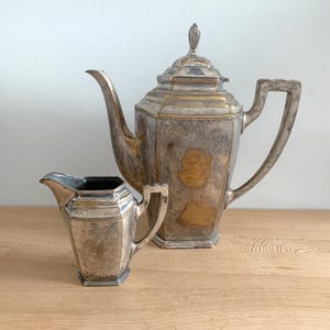 May include: A silver and gold teapot and creamer set. The teapot has a geometric design and a gold accent on the lid. The creamer is a smaller version of the teapot with a similar design.