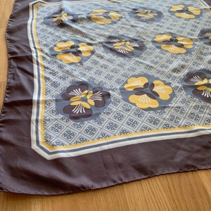 May include: A gray silk scarf with a white and yellow floral pattern. The flowers are stylized and have a geometric design. The scarf has a white border with a yellow stripe.
