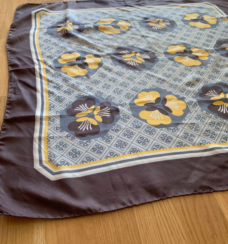 May include: A gray silk scarf with a white and yellow floral pattern. The flowers are stylized and have a geometric design. The scarf has a white border with a yellow stripe.