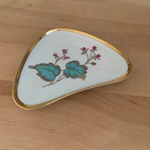 May include: A white ceramic dish with a gold rim and a floral design. The design features teal leaves and pink flowers.