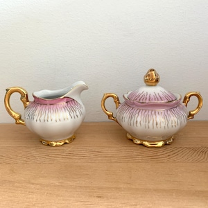 Peut inclure: Un ensemble de crémier et de sucrier en porcelaine blanche avec des accents roses et dorés. Le crémier et le sucrier ont un motif floral délicat avec des bordures dorées.