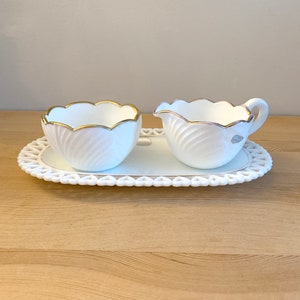 Könnte beinhalten: Weißes Porzellan-Zucker- und Sahne-Set mit goldener Verzierung auf einem weißen Tablett mit Spitzenrand.
