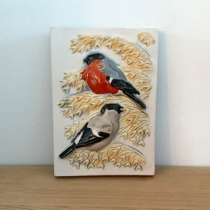 以下が含まれることがあります： 小麦の穂先に止まっている2羽の鳥の3Dデザインが施された白いセラミックタイル。鳥はグレー、ブラック、レッドの色合いで描かれています。