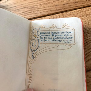 May include: An open book with Swedish text, showcasing detailed calligraphy and decorative elements in blue, gold, and brown. The text reads "jungen till herrens &auml;ra, loven hans namn, f&ouml;rkunnen ifr&aring;n dag till dag gl&auml;djebudskapet om hans fr&auml;lsning. Ps. 96:2."