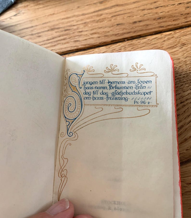 May include: An open book with Swedish text, showcasing detailed calligraphy and decorative elements in blue, gold, and brown. The text reads "jungen till herrens &auml;ra, loven hans namn, f&ouml;rkunnen ifr&aring;n dag till dag gl&auml;djebudskapet om hans fr&auml;lsning. Ps. 96:2."