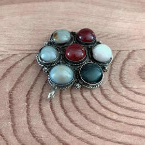 Puede incluir: Broche plateado con dise&ntilde;o floral. El broche presenta siete gemas redondas y coloridas en tonos rojo, blanco, verde y azul. Las gemas est&aacute;n engastadas en biseles individuales y dispuestas alrededor de un punto central.