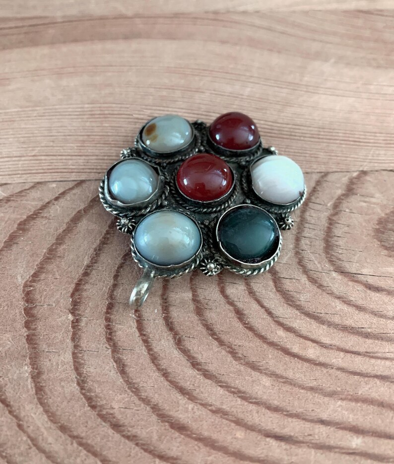 Puede incluir: Broche plateado con dise&ntilde;o floral. El broche presenta siete gemas redondas y coloridas en tonos rojo, blanco, verde y azul. Las gemas est&aacute;n engastadas en biseles individuales y dispuestas alrededor de un punto central.