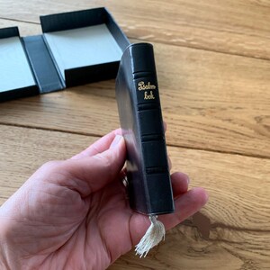 May include: A miniature black book with gold lettering reading "Psalm bok." It features a tassel and is held in a hand. A black box with a white interior is visible in the background.