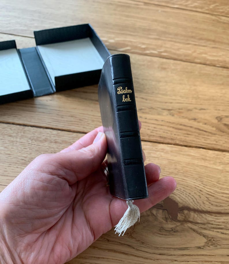 May include: A miniature black book with gold lettering reading "Psalm bok." It features a tassel and is held in a hand. A black box with a white interior is visible in the background.