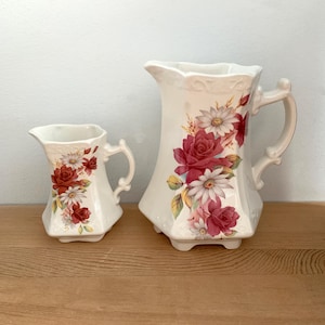May include: A set of two white ceramic jugs with floral designs. The larger jug has a wide mouth and a handle, while the smaller jug has a narrower mouth and a handle. Both jugs feature a floral pattern with red roses, white daisies, and green leaves.