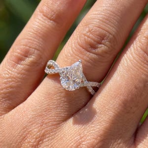 Puede incluir: Un anillo de tono plateado con una piedra central en forma de pera y una banda dividida y retorcida incrustada con pequeñas piedras transparentes en pavé. La banda de metal tiene un acabado pulido y la piedra central se sujeta con un engaste de garras.