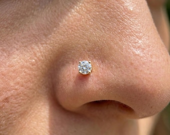 Puede incluir: Primer plano de un piercing de nariz dorado con una gema redonda y transparente. El perno está engastado en una base dorada y está posicionado en el costado de una nariz. Se ve la piel alrededor de la nariz.