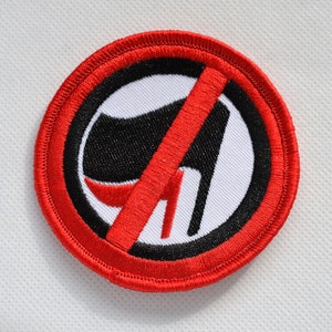 Puede incluir: Un círculo rojo con un contorno negro y una línea diagonal roja que atraviesa una bota negra con un tacón rojo. La bota está sobre un fondo blanco.