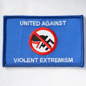 Könnte beinhalten: Ein blauer Patch mit weißen Nähten an den Rändern. Der Patch trägt den Text "UNITED AGAINST" oben und "VIOLENT EXTREMISM" unten. In der Mitte befindet sich ein roter Kreis mit einem diagonalen Strich durch ihn. Im Kreis befindet sich ein schwarz-weißes Symbol einer Faust mit einem Hakenkreuz.