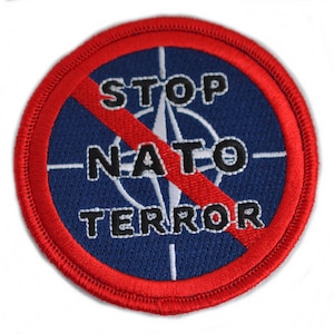 Könnte beinhalten: Ein roter Kreis mit einer diagonalen roten Linie durch ihn. Der Kreis hat einen blauen Hintergrund mit einer weißen Windrose und dem Text "STOP NATO TERROR" in Schwarz.