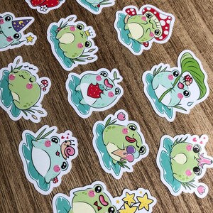 May include: A set of 11 frog stickers with various designs. The frogs are green and have different expressions and accessories, such as a crown, a strawberry, and an ice cream cone. The stickers are on a light brown wooden surface.