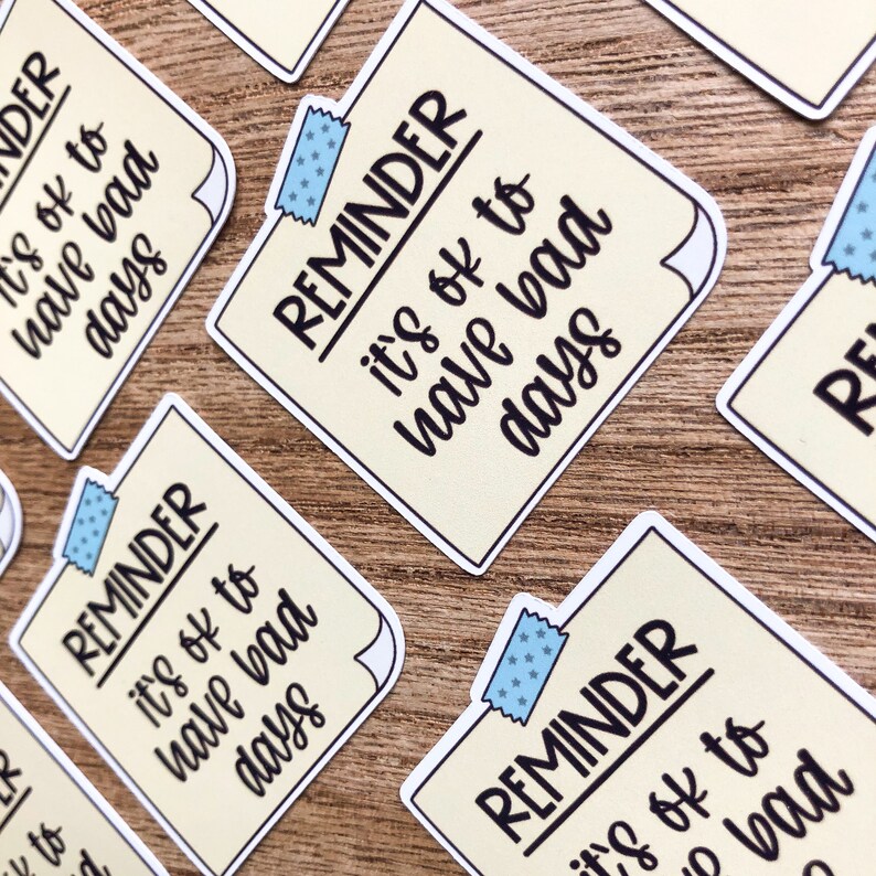 Puede incluir: Un conjunto de cuatro pegatinas de recordatorio amarillas con acentos de cinta azul. Cada pegatina presenta el texto "REMINDER it's ok to have bad days".