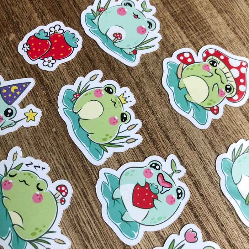 May include: A set of nine frog stickers with various designs. The stickers feature frogs in different poses, including sleeping, eating strawberries, wearing a crown, and holding a mushroom. The stickers are on a wooden background.