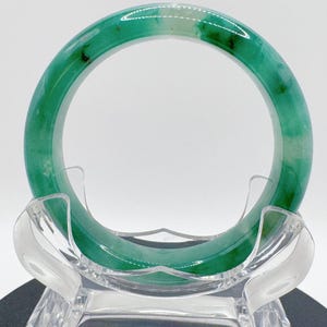 May include: A green jade bangle bracelet. The polished stone shows a mix of light and dark green shades, with subtle colour variations. The bracelet is displayed on a clear acrylic stand, highlighting its smooth, circular form.