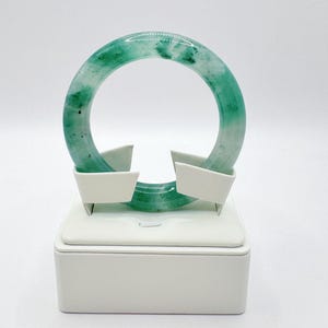 May include: A jade bangle bracelet, predominantly green with white marbling, is displayed on a white stand. The bracelet is a complete circle, showcasing the natural variations in the stone's color and texture. The stand is a simple, rectangular design.