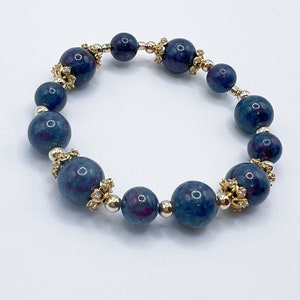 Op de afbeelding: Een blauwe en paarse edelsteen armband met gouden accenten en kleine heldere stenen. De armband is gemaakt van ronde kralen.