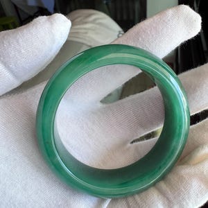 May include: A translucent green jade bangle bracelet. The jade is a rich, deep green color with subtle variations in tone. The bracelet is circular and smooth, reflecting light. A classic piece of jewelry.