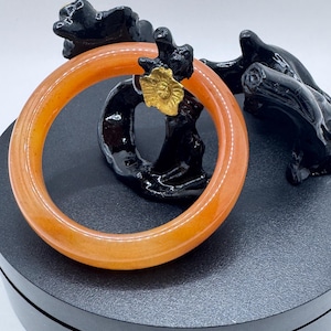 Könnte beinhalten: Ein glatter, durchscheinender orangefarbener Armreif mit einem kleinen, strukturierten goldfarbenen Blumenakzent an der Oberseite. Der kreisförmige Armreif hat eine polierte Oberfläche und ist an einer dunklen, abstrakten Skulptur angelehnt.