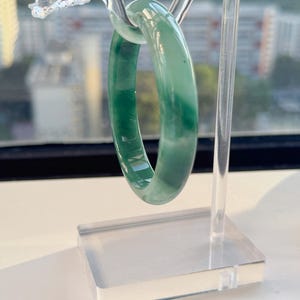 May include: A jadeite bangle bracelet in shades of green is displayed on a clear acrylic stand. The stand features a dragon head design at the top. The bracelet is approximately 2.5 inches in diameter.