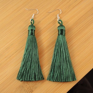 May include: A pair of dark green tassel earrings with silver-coloured hooks. The tassels are made of silky threads, displayed on a light brown wooden surface.