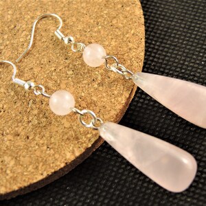 May include: A pair of earrings featuring a silver hook, a small round pink bead, and a larger teardrop-shaped pink stone. The earrings are displayed on a cork surface.