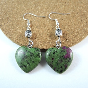 May include: A pair of heart-shaped earrings. Each earring has a green stone with dark red and black inclusions, suspended from a silver-coloured hook and decorative bead. The earrings are displayed on a light brown surface.