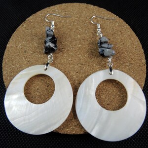 May include: Earrings with large, white, circular pendants and a central hole. Each earring has a silver hook and a cluster of black and grey speckled stones. The earrings are displayed on a brown, textured surface.