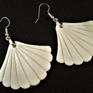 Könnte beinhalten: Ein Paar cremefarbene, fächerförmige Ohrringe mit silberfarbenen Haken. Die Ohrringe haben einen gewellten Rand und parallele Linien, die von oben ausstrahlen und ein strukturiertes Aussehen erzeugen. Der Hintergrund ist schwarz.