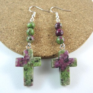 Può includere: Orecchini con pendenti a forma di croce in pietra screziata verde e magenta. Le croci sono appese a ganci color argento e sono decorate con perline verdi e magenta.