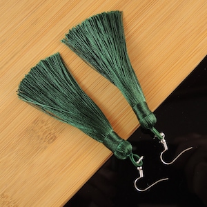 May include: A pair of dark green tassel earrings with silver fish hook ear wires. The earrings feature silky threads and are displayed on a light brown and black surface.