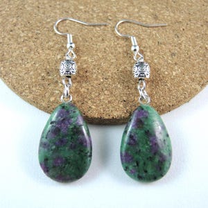 May include: Dangle earrings with teardrop-shaped green and purple zoisite stones. The earrings have silver-coloured hooks and decorative beads. The stones are polished, showing a natural, mottled pattern.