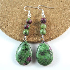 May include: A pair of dangle earrings featuring green and red gemstone beads. The earrings have a silver-colored hook and are composed of a series of small, round beads leading to a teardrop-shaped stone. The teardrop stones are green with red and black inclusions.